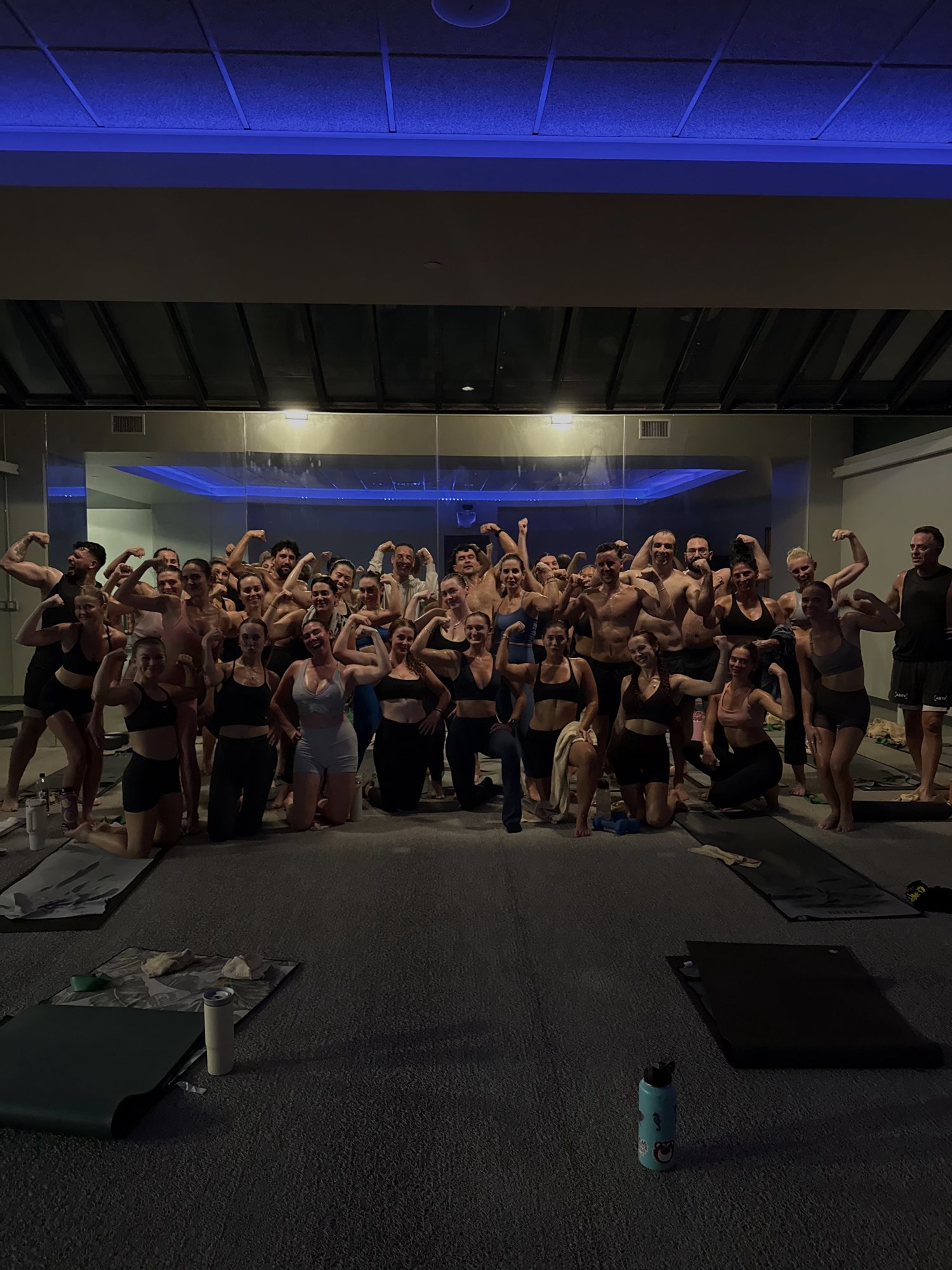 A large group flexing their arms together in a dimly lit studio with blue ceiling lights